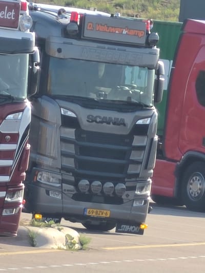 A close-up shot shows the front of a grey Scania truck parked next to a red truck. The grey truck's grill and headlights are visible. Text on the grey truck includes "INTERCOOLER" and "SCANIA". A yellow license plate reads "69-BZV-6". A small orange indicator light is illuminated above the license plate. Text on a sign on the left side of the grey truck reads "D. Veluwenkamp". The scene is outdoors, possibly a truck stop or industrial area, with a glimpse of greenery and a paved surface. The image appears to be taken during daylight.