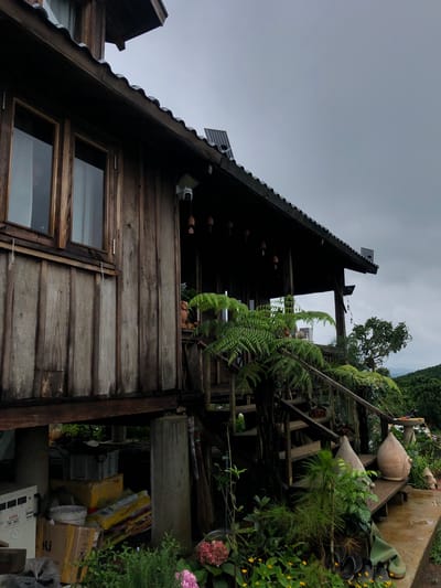This is a medium-angle, eye-level exterior shot of a weathered wooden stilt house on a cloudy day in Thôn 4, Vietnam. The house features dark, corrugated roofing and is constructed from vertical wooden planks. To the left, a window with wooden frames is visible. A security camera is mounted on the wall above a porch area. Hanging from the eaves are small, decorative bells. Lush green ferns and other plants grow on and around the wooden porch and staircase leading up to the house. To the right, a large, urn-like ceramic pot sits on the porch, with a landscape of lush green hills and trees visible in the background. The ground level beneath the house is cluttered with various items, including boxes, a bucket, and what appears to be a broom. There are no people visible in the image.