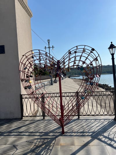 A large red metal heart sculpture, adorned with numerous padlocks, stands prominently in the foreground. The sculpture is located outdoors, adjacent to a promenade or walkway that runs alongside a body of water. To the left of the heart is a light-colored, textured wall of a building. A black metal fence runs in front of the sculpture and separates it from the promenade. A lamppost with a star on top is visible behind the sculpture, and another, more traditional lamppost is to the right. The background features a body of water, hills with trees, and a clear blue sky. The scene is illuminated by sunlight, casting long shadows from the sculpture and fence onto the tiled ground. The city of Korzhevo, Moldova is mentioned in the prompt.