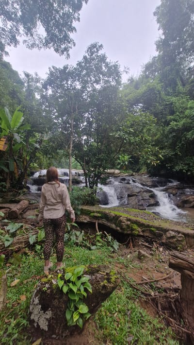 A woman stands with her back to the camera, facing a cascading waterfall in a lush, green jungle environment. She is wearing a light brown collared shirt and leopard-print leggings. The waterfall is rocky and multi-tiered, with white water rushing over the dark stones. Tall trees and dense foliage surround the waterfall, creating a dense canopy overhead. The ground in the foreground is covered with moss, grass, and fallen logs. The overall scene depicts a natural and somewhat wild landscape, likely in a tropical region. The location is identified as Pong Yaeng, Thailand.