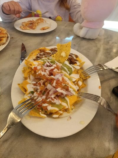 A close-up captures a plate of loaded nachos featuring cheese, guacamole, pico de gallo, sour cream, and a lone tortilla chip. Three forks are poised to dig in. Another plate contains pizza slices. A knife lies next to the plates. In the background, a person in a long-sleeved shirt and a stuffed animal are visible at the table. The surface appears to be a marble tabletop.