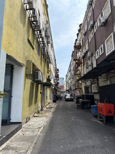 An urban alleyway bisects rows of tall buildings adorned with numerous air conditioning units. One building has a light yellow facade, while the building opposite it is brown and contains a fire escape. A dark, narrow canopy stretches above the alley. A silver car and a blue barrel are also visible along the asphalt-covered road. The sky is light blue, indicating daylight. The overall scene is a densely populated urban environment.