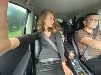 In the city of Sepych, Russia, this image captures a frontal view from inside a vehicle's passenger seat, looking towards the driver and the road ahead. The driver, a male with a close-cropped haircut and glasses, wears a brown collared shirt and a seatbelt. Next to him, in the passenger seat, a woman with shoulder-length brown hair wears a black long-sleeved dress and a seatbelt. Her left arm is extended towards the camera, obscuring part of the frame. In the background, a child is visible in a car seat. The car's interior features black leather seats and a center console with a bottle, a phone, and buttons. Outside the car windows, lush green trees line the road, suggesting a rural or suburban setting. The sky appears overcast, and the window is speckled with raindrops, indicating wet weather.