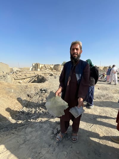 The image captures a man standing in a scene of extensive structural damage in Spin Boldak, Afghanistan, under a clear blue sky. The central figure, a man with a beard, wears a brown long-sleeved garment, a dark vest, and a light-colored cap, along with sandals. He holds two irregular fragments of debris: a larger, light grey-green piece in his left hand and a smaller, dark grey piece in his right. He faces the camera with a somber expression.
In the mid-ground to the right, several other individuals are visible. One person is partially obscured by a dark green head covering. Further right, a group of men in traditional attire stands amidst the rubble.
The background reveals a landscape of collapsed and severely damaged buildings, characterized by piles of bricks, concrete, and various structural wreckage. The ground is dusty, uneven, and covered with debris. Shadows suggest bright, direct sunlight. A utility pole is visible in the distant right background.