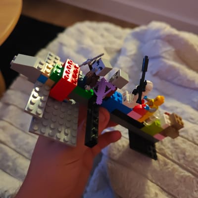 A hand is holding a Lego creation that appears to be a vehicle with figures on it. The vehicle is constructed from various colored Lego bricks including grey, red, green, blue, and yellow. There are at least three figures: two with grey, block-like heads and brown and purple bodies, and one with a light skin tone, yellow body and blue hands. One of the grey-headed figures appears to be holding a weapon. The Lego creation is positioned on a white, fluffy surface, possibly a blanket or couch. The background is blurred, suggesting a shallow depth of field. The scene does not contain any discernible landmarks or features that would indicate a location in Locking Parklands, United Kingdom.