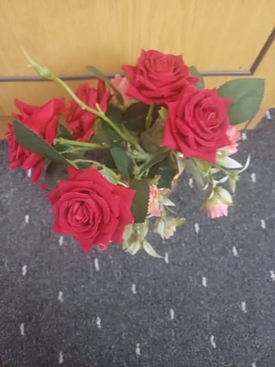 The image shows a close-up, high-angle shot of a bouquet of artificial red roses and some pink buds, arranged in a vase which is mostly out of frame at the bottom. The flowers have green leaves and stems. The bouquet is placed on a dark grey carpet with a subtle pattern of small white speckles. In the background, a light yellow-brown wooden surface is visible, likely a wall or furniture. The scene appears to be indoors, possibly in a domestic setting. There are no people or animals present. The lighting is moderate. This image is not specific to Jumashuy, Uzbekistan, and does not provide any identifiable landmarks or cultural indicators of that location.