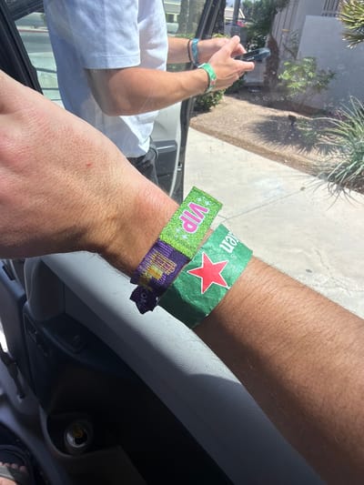 A close-up, brightly lit, daytime shot shows two human arms inside a vehicle, possibly a car, given the dashboard visible in the lower right corner. The arm in the foreground has a bright green, glittery VIP wristband and beneath it, a wristband that reads "COACH." The other arm further away from the camera has a green wristband with a large red star. The arms are positioned close to the windshield. Outside the windshield, a slightly blurred scene depicts palm trees and foliage, indicating an outdoor setting. Judging from the "COACH" wristband it is very likely this is a photo taken in the Coachella valley, in Palm Desert United States of America. Another arm is visible farther away holding what appears to be a cell phone. The arm with the wristbands appears to be resting on the door frame and the other on the driver's steering wheel.