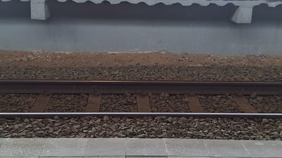 The image shows a section of train tracks in Karawang, Indonesia. The tracks consist of two parallel metal rails resting on wooden sleepers, which are embedded in a bed of gravel. Above the tracks, there is a concrete wall and overhang. The foreground shows a paved area. There are no people visible in the image.