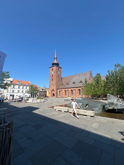 The image shows a bright, sunny day in a town square in Horsens, Denmark. A young boy in a white t-shirt and beige shorts is walking in the foreground, with his left hand outstretched. Behind him is a large, modern fountain with dark, sculpted metal elements. In the background, a tall, red brick church with a steeple stands prominently. The church has a clock face visible on its tower. To the left of the church, there are several buildings with red and white facades, some with shops. The ground of the square is paved with large, dark gray tiles. Trees are visible to the right of the church, and a few people are seen in the distance walking on the sidewalk in front of the buildings. There is a shadow cast across the square from an unseen object or structure, creating a strong contrast between light and dark areas. A white banner is partially visible on the left side of the image.