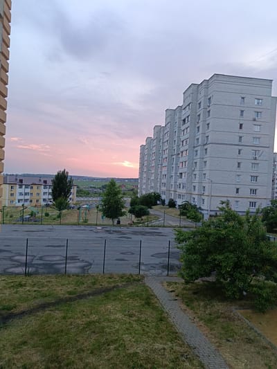 The image depicts an outdoor scene in Gubkin, Russia, during dusk. The sky is a soft gradient of pink and purple hues, indicating the sun has recently set. A long, multi-story apartment building with a light-colored brick facade dominates the right side of the frame. Its numerous windows, some with air conditioning units, are visible. To the left of this building, a slightly shorter, yellow and white apartment building with a brown roof is partially visible, showing balconies and windows.

In the foreground, a paved area with cracks and dark patches resembling puddles or stains is bordered by a black metal fence. Beyond the fence, a grassy area transitions into a courtyard with playground equipment, including swings and a small shelter with a picnic table. Two trees, one tall and dark green and another shorter and lighter green, stand in the courtyard. Further back, a road is visible, with a few parked cars and more residential buildings in the distance, nestled amongst rolling hills. A lone figure is seated on a bench near the playground, appearing to be observing the surroundings. The overall atmosphere suggests a quiet evening in a residential urban area. No visible text is present in the image.