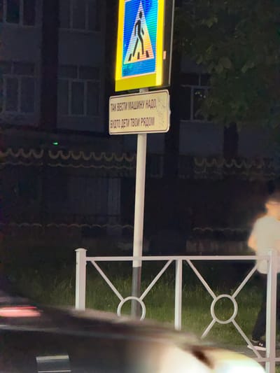 This image depicts a nighttime scene on a street in Novokuli, Russia. A pedestrian crossing sign, featuring a blue triangle with a white silhouette of a person walking and yellow border, is prominently displayed. Below the pedestrian sign is a rectangular sign with black text on a yellow background. The text reads, "ТАК ВЕСТИ МАШИНУ НАДО, БУДТО ДЕТИ ТВОИ РЯДОМ." This translates to "Drive the car like your children are nearby." The sign also has small cartoonish drawings of children.

In the background, a building with windows is visible. A white metal fence with a decorative pattern of intersecting lines and circles runs horizontally across the lower portion of the image. Grass is visible behind the fence. A blurry figure of a person wearing dark pants and shoes is partially visible to the right, standing near the fence. Another blurry figure, possibly in the foreground, obscures part of the scene. The lighting suggests it is nighttime, with artificial lights illuminating the area. The overall impression is a residential or pedestrian-heavy area where safety is emphasized through signage.