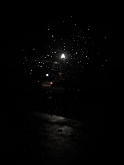 This image depicts a nighttime scene observed through a rain-streaked transparent surface, likely a window or windshield, in the city of Griboyedov, Armenia. Numerous water droplets of varying sizes are scattered across the foreground, causing diffraction and blurring of light sources. In the upper-center, a prominent, elongated white light source, consistent with a streetlamp, is visible. To its lower-left, a smaller, circular white light appears. Further back and slightly below this second light, a small, circular orange or red light is present. The lower-center reveals an indistinct, reflective greyish-white area on what appears to be a wet ground surface, possibly a street or pavement, reflecting ambient light. The remainder of the background is predominantly dark and featureless, indicating low light conditions and lack of specific detail. No individuals or specific objects are clearly discernible beyond the light sources and wet surfaces.