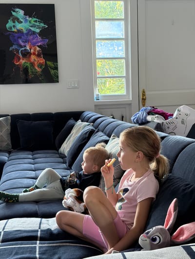Two children are seated on a large, navy blue sectional sofa in a living room. The child on the left is a toddler with blonde hair, wearing a black t-shirt and light grey pants with colorful socks. The toddler is lying down, holding a stuffed animal. The child on the right is a pre-teen girl with blonde hair pulled into a ponytail, wearing a pink t-shirt and pink shorts. She is sitting upright, looking to the left and eating a snack. A stuffed bunny, possibly Judy Hopps from Zootopia, is positioned next to her. The sofa is adorned with several decorative pillows. Behind the sofa, a window with multiple panes offers a view of greenery and sky. On the wall to the left of the window, an abstract artwork with vibrant colors is displayed. To the right of the window is a closed door with a golden doorknob. A stack of folded clothes and some paper bags are visible on the back of the sofa, behind the girl. A plaid blanket is draped over the lower section of the sofa. The room appears to be well-lit, with natural light streaming in from the window. The scene suggests a casual, indoor activity, likely watching television or relaxing. This location is not within the city of Flanders, United States of America; the image appears to be located in a different country and city.