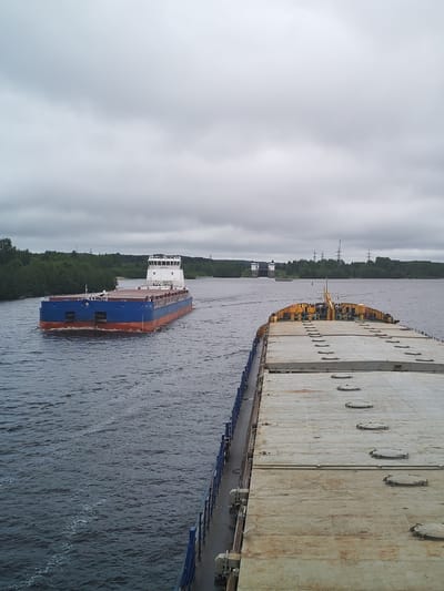 The image depicts a wide river landscape with two large barges navigating the water. In the foreground, the deck of a barge is visible, with a faded gray surface featuring numerous access hatches. Yellow structures are visible near the rear of the barge. To the left, another barge is moving in the opposite direction. It is painted with a red and blue hull and has a white, multi-story structure at the top. In the distance, a structure with a tall white tower is visible, possibly a lock or dam. The landscape includes green trees lining the riverbanks and utility towers visible on the shoreline. The sky is overcast with heavy clouds. The location is in the city of Анхимово, Russia.