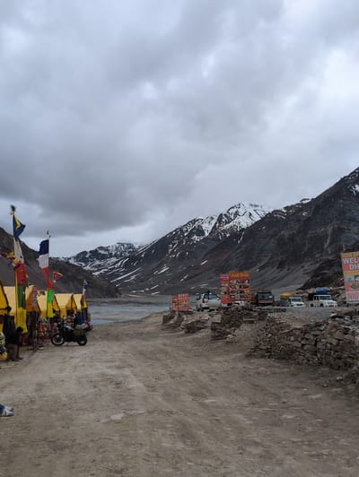 The image depicts a roadside scene in Killingsarai, India, with a backdrop of majestic, snow-capped mountains under a cloudy sky. In the foreground, a dirt road leads to a cluster of yellow tents, likely temporary stalls selling goods, adorned with colorful flags. A motorcycle is parked near the stalls, suggesting a mode of transport for travelers. Further down the road, several vehicles, including a white van, a truck, and two white SUVs, are parked. Prominently displayed are large signboards advertising "HIMAL PUNJABI DHABA" and "NEW BHAI DHABA," indicating food establishments. The scene is framed by rough, stone-built walls on the right side, adding to the rugged, mountainous terrain. The environment suggests a remote location along a mountain pass, possibly a resting point for tourists or travelers.