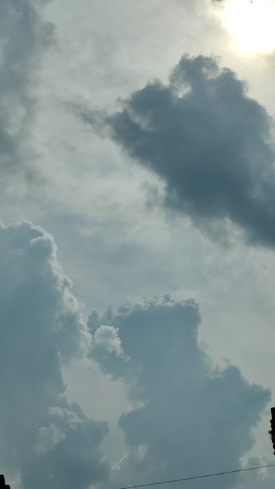 The image captures a cloudy sky over Bolotnoye, Russia. The sun is partially obscured by thick, dark gray clouds, casting a diffused light. Several large, fluffy white and gray clouds dominate the frame, with some appearing almost solid and others wispy and translucent. The lower portion of the image shows a glimpse of what appears to be a dark, silhouetted structure on the right edge, possibly a building or pole, with a thin wire running horizontally across the bottom.