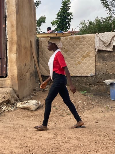 The scene depicts an unpaved outdoor area in Idiroko, Nigeria, under an overcast sky. In the foreground, a young woman, appearing to be of African descent, walks from left to right in profile. She wears a red short-sleeved shirt with a ruffled hem, black skinny jeans, and brown leather flip-flops. A light-colored cloth is wrapped around her head, and she balances a light-colored, possibly metal, shallow basin or tray containing indistinct items on her head. Her arms swing naturally with her stride.
To the left, a weathered, light brown building wall with visible text or graffiti leans against a corrugated metal panel. A wooden plank stands vertically against this wall. Near the base of the wall, a white sack and debris are visible on the dirt ground. In the background to the right, a large, worn, quilted mattress is propped or hung against a dark, possibly block, wall. An additional piece of fabric is draped over the top right corner of the mattress. Behind these structures, dense green foliage and trees are visible. A small child's head is partially visible above a distant structure, to the left of the prominent tree. On the far right, a blue plastic bucket rests on the ground. The ground throughout the image is composed of reddish-brown dirt or sand.