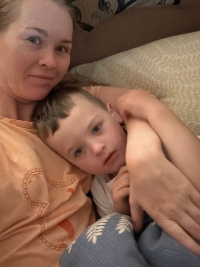 This is a close-up, overhead shot of a mother and child lying down together. The mother's face is on the left side of the frame, with her eyes open and looking towards the camera. Her hair is blonde and appears to be pulled back. The child is positioned above her, with their head resting on her chest and arm. The child has light brown hair and blue eyes and is looking directly at the camera with a calm expression. They are both wearing casual clothing. The child is wearing a light-colored t-shirt, and the mother is wearing an orange shirt with a decorative pattern. They are lying on a bed with light-colored bedding and a patterned blanket. The background suggests an indoor setting, possibly a bedroom, with soft lighting. The overall mood is one of intimacy and comfort.