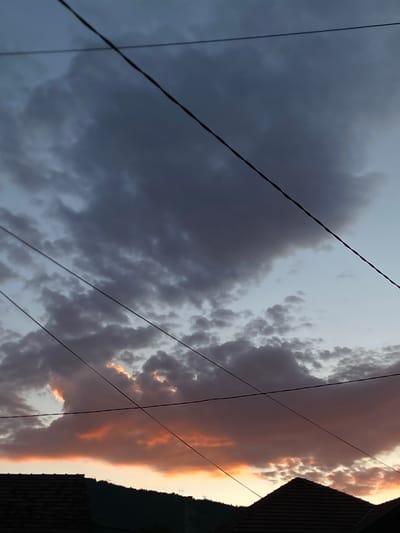 The image depicts a cloudy sky with hues of dark gray, white, and orange, suggesting either sunrise or sunset. Multiple dark electrical wires traverse the sky diagonally. Silhouetted against the sky are the dark roofs of buildings in the foreground and a tree-covered hillside in the background. The scene appears to be set in a residential area, possibly within the city of Prijepolje Municipality, Serbia, given the architecture and the overall atmosphere. There are no people or animals visible in the frame. The lighting is dim, consistent with twilight conditions.