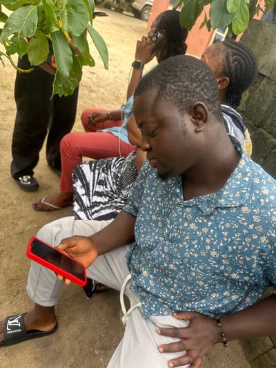 In Ganyingbo, Nigeria, an outdoor scene shows a man in the foreground holding a red smartphone and wearing a blue floral shirt and gray pants. He is seated on the ground, with Louis Vuitton sandals visible on his left foot. Behind him, two women are seated, one wearing red pants and a black and white striped dress, and the other with braided hair, speaking on a phone. Another person, partially visible, stands to the left wearing black pants and shoes. Green leaves from a tree branch hang over the scene, and a textured wall is present on the right. The ground appears to be a mix of sand and dirt.