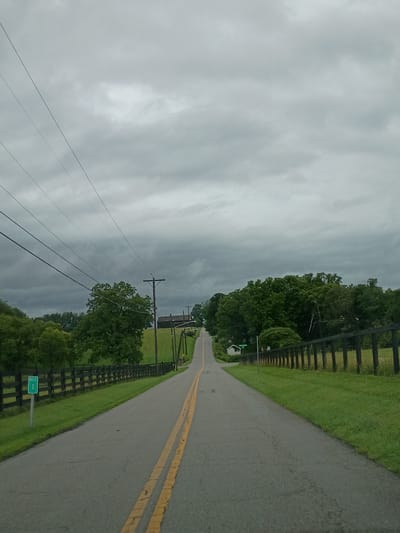 The image depicts a rural road scene under a cloudy sky. The road is paved and has double yellow lines. On the left side of the road, there is a black wooden fence bordering a grassy area. A green sign with "MILE 1" in white is visible near the fence. Beyond the fence, there are trees and utility poles with wires. On the right side of the road, there is also a black wooden fence and a grassy area, with trees in the distance. The road recedes into the distance, and there are some buildings and more trees visible further up the road. The environment appears to be a countryside setting. There are no people or animals visible in the image.