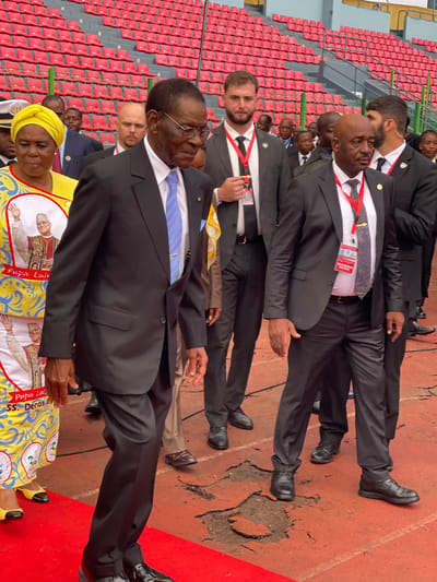 A low-angle, canted photograph captures a scene featuring a group of people in dark suits. A man in the front is wearing glasses, a blue tie, and a dark suit. Beside him is a woman in a yellow patterned headscarf and dress with images of a man on it, and a white bib. The individuals are lined up, possibly standing on a cracked surface. There are red bleacher seats in the background. The location appears to be a stadium in Malabo, Equatorial Guinea, though this can't be confirmed with the image. The overall impression is one of formality or ceremony.