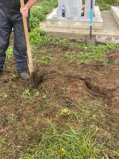 The image shows a gravesite in Makariopolsko, Bulgaria. A person is digging with a shovel in the ground near the headstone. Another shovel is leaning against the headstone. A bottle of soda is also visible near the headstone. The ground is covered with grass and dirt.