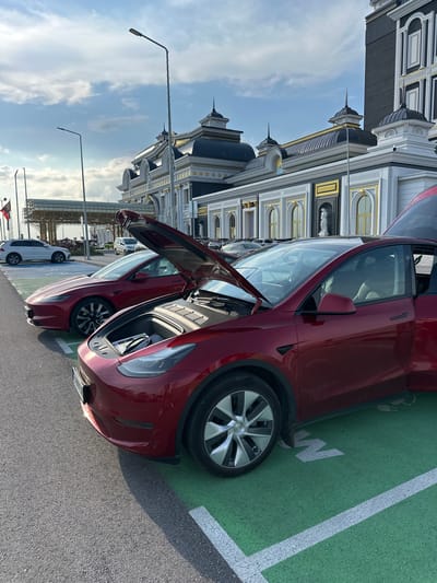 The image depicts an outdoor parking lot in Kapitan Andreevo, Bulgaria, featuring two red Tesla electric vehicles. In the foreground, a red Tesla Model Y is parked in a green-marked electric vehicle charging bay, indicated by a white charging symbol and a partial white 'M' or 'W' on the green pavement. The vehicle's front trunk (frunk) is open, revealing a storage compartment containing a dark, rectangular object resembling a folder or manual. The driver's side door is also open. Directly behind the Model Y, a red Tesla Model 3 is parked with its hood open.
The background features a large, ornate building exhibiting classical architecture, characterized by dark grey/black domed roofs, white facade elements, gold trim, arched windows, and decorative cornices. A white statue is visible in a niche on the building's facade. Further to the left, a white SUV is parked, and a covered walkway structure with metal framework is visible. Several tall streetlights stand throughout the scene. The sky is partially cloudy, and sunlight casts shadows on the asphalt, indicating daytime. No people are visible in the frame.