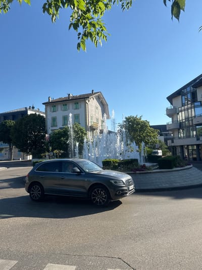 A dark gray Audi SUV is captured in motion on a paved road in Divonne-les-Bains, France. The vehicle is positioned in the foreground, on the right side of the frame, partially obscuring a circular fountain with multiple water jets. Behind the fountain, several buildings are visible. To the left of the fountain stands a light-colored, multi-story building with green shutters and balconies. Further to the left, another building with a dark roof is partially visible, and a cluster of trees is situated between the two buildings. To the right of the fountain, a modern building with a glass facade and balconies is present. The scene is illuminated by bright sunlight, casting shadows on the road. The sky is clear and blue, with the leaves of a tree hanging into the upper portion of the frame. A speed limit sign reading "ZONE 30" is visible near the fountain.