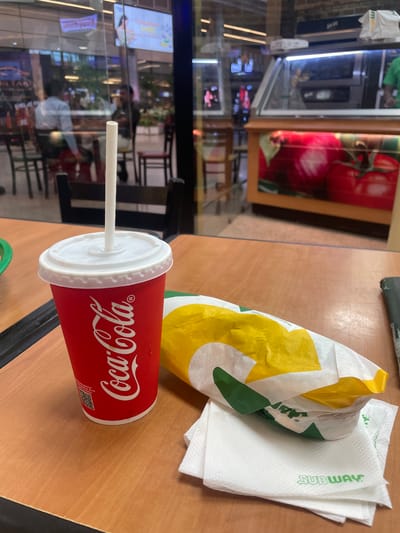 An overhead shot shows a Subway sandwich and a Coca-Cola cup on a wooden table. The Coca-Cola cup is red with a white lid and straw. The sandwich is wrapped in yellow and white paper with green accents. The Subway logo is visible on a white napkin in front of the sandwich. In the background, people are seated at tables inside what appears to be a food court, possibly at an airport or mall in Gollapalle Khurd, India, as indicated by the signage visible through the glass partition. A display case with images of tomatoes is also visible.