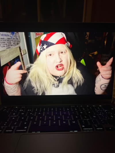 This media file captures an image displayed on a laptop screen, with the laptop's keyboard visible on the right side. The scene is set in Torrent, Spain.
The main subject is a person, likely young, with very pale skin, bright blonde hair, and reddish-brown eyes, characteristics often associated with albinism. They are wearing a bandana or head covering with an American flag design. The person has several facial piercings around their nose and mouth, and their mouth is open, revealing teeth. They wear a dark top and a dark, possibly spiked, collar or necklace.
Tattoos are visible on both of the person's arms. On their left arm (viewer's right), a Yin-Yang symbol and other abstract designs are apparent, along with a purple beaded bracelet. On their right arm (viewer's left), script-like text is visible, including partial words like "Death totally bite" and what appears to be "FUCK," along with a red string bracelet. One hand is raised, fingers spread, while the other is partially visible, seemingly making a gesture.
Below the laptop screen, a document or paper is visible with text, including "MERICANA INDEPENDENTE DELA" and "YUCA." The laptop's keyboard is backlit, suggesting a dim environment, and features typical MacBook keys such as "option," "command," and "esc," along with letters and numbers. The image appears to be a photograph taken of the screen, rather than a direct digital capture.