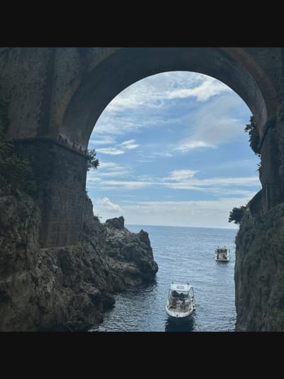 This image shows a view through a large stone archway, likely part of a bridge or aqueduct, looking out at the sea. Two white motorboats are visible on the water. The left side of the archway is lined with rough-hewn stone blocks, with some vegetation growing on the rocky cliffs. The right side of the archway features a stone structure with visible stairs, also surrounded by greenery. The sky is blue with scattered white clouds. The scene depicts a coastal landscape with a significant architectural element. The location is identified as Norcia, Italy.