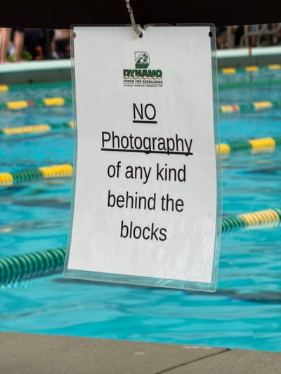 An indoor swimming pool facility in Chamblee, United States of America, is depicted with a white sign in a clear plastic holder as the central focus. The sign hangs from a dark, blurry overhead structure by a silver metal hook attached to a small metal ring punched through the top left corner of the plastic holder. The sign displays a green and white logo featuring a mountain peak with a "D" inside, over intertwined rings, with "DYNAMO CODED FOR EXCELLENCE" printed below it, followed by "ATHLETICS • ACADEMICS • FRIENDSHIPS • FUN!" in smaller text. The primary message, printed in bold black capital letters, states: "NO Photography of any kind behind the blocks." "NO" and "Photography" are underlined. The background features the vibrant turquoise water of a swimming pool, traversed by numerous lane lines in yellow and green/white. The water surface shows subtle ripples. Blurred figures, visible mainly as legs and lower torsos, are present on what appears to be a pool deck or near diving blocks in the distant background. The foreground shows a slightly blurred edge of a light gray concrete or tiled surface.