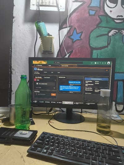 The scene is an interior shot of a workspace. It appears to be in a residential or home office setting in Nepanagar, India (21.45, 76.397). On a wooden desk, a Lenovo computer monitor is displaying a program or software with a chat interaction window. In the chat window, a message reads, "Hello, thank you for contacting Intrepid. I am Anna, and I would be delighted to help you." The user appears to have entered a message, as there is an "Enter message" box at the top of the screen, which is partially obscured. There is a green glass bottle, partially filled with a liquid, and a glass on the desk to the left of the monitor. A black keyboard is positioned below the monitor. On the right side of the desk, a clear glass bottle filled with a yellowish liquid sits on the desk. The wall behind the desk is painted in a bright, colorful abstract design, possibly representing a cartoon character or a stylized representation of a person. A comb rests in a plastic cup on the wall. The desk is uncluttered, with only the computer, keyboard, glass bottle, and glass present. The scene appears to depict a work-from-home setup.