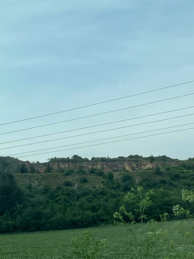 The image presents a vertical view of a landscape scene in or around Ushintsi, Bulgaria. A hillside with a rock face and abundant green vegetation dominates the right side of the frame. Power lines run diagonally from the top left to the center, implying a nearby settlement. The sky is overcast and light blue. To the right of the hillside is an open field with green vegetation. There are no people visible.