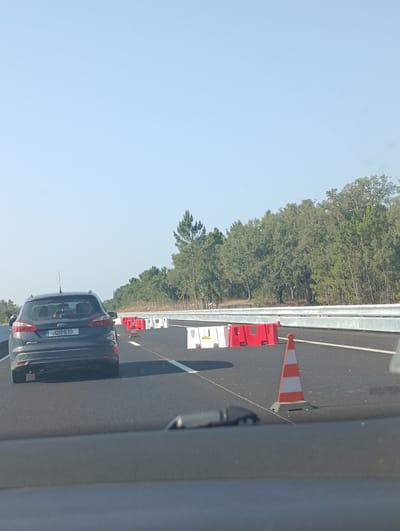 A dark gray Ford Focus is driving on a highway in Santiago do Cacém, Portugal. The car is facing away from the camera, and its license plate reads "01MI19". The highway is lined with orange and white traffic cones and temporary white and red barrier walls. Trees and foliage line the side of the highway. The sky is clear and blue. The image appears to be taken from inside another vehicle.