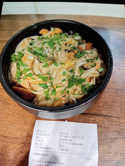 A close-up, overhead shot shows a black bowl filled with noodles, seafood, and greens, placed on a wooden table. A receipt is partially visible in the bottom right corner of the frame. The receipt appears to be from a restaurant in Mykolaiv, Ukraine, with a date of June 10, 2026, and a check number of 36081. The food item listed is "Локшина з Морепродуктaми" which translates to "Noodles with Seafood."