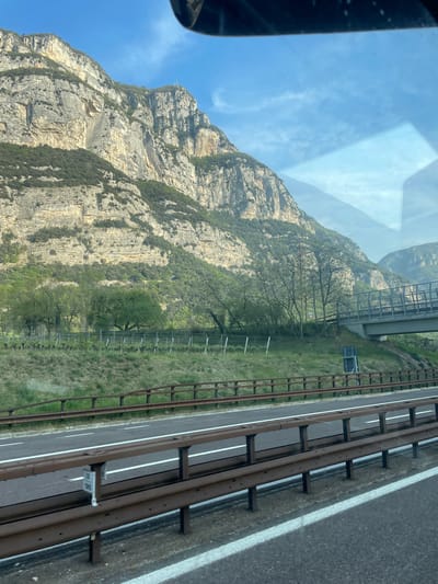 The image shows a view from a vehicle moving along a highway in Brentino, Italy. A large, rocky mountain dominates the left side of the frame, its terrain marked by layers of exposed rock and sparse vegetation. To the right, a highway is visible, separated from the adjacent terrain by a guardrail. A bridge spans the area between the mountain and the highway. The sky, visible through the windshield, is partially obscured by reflections.