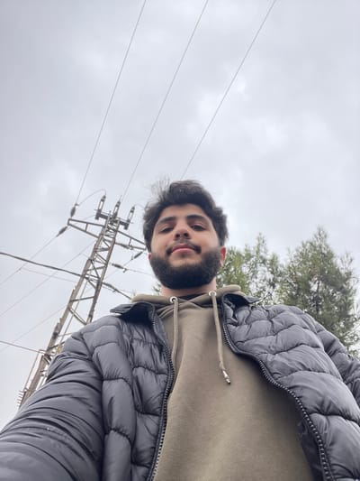 A low-angle, wide-shot image depicts a male subject looking directly at the camera in Dahiyat Harasta, Syria, under an overcast sky. The subject, positioned centrally, is a young adult male with dark hair and a beard, wearing an unzipped black puffer jacket over an olive green hooded sweatshirt. His expression is neutral to slightly smiling. Behind the subject, to the left, a metal electricity pylon with multiple insulators and power lines is visible, extending upwards towards the top left of the frame. To the right, a portion of a conifer tree is visible. The sky is uniformly gray and cloudy, occupying the upper two-thirds of the image.