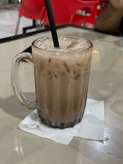 This image, captured in Lhok Mereubo, Indonesia, depicts a clear glass mug containing an iced, light brown beverage with a black plastic straw. The mug features a handle on its left side and a vertically ribbed or faceted lower half. It rests on a folded white paper napkin or coaster, which is positioned on a smooth, light-colored tabletop. In the blurred background, partially visible red chairs indicate a seating area, and a dark, indistinct form to the right suggests the presence of a person or a dark object on the table.