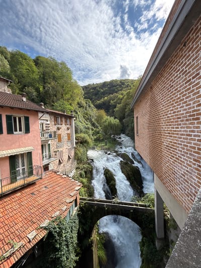 The image depicts a panoramic view of the Orrido di Nesso waterfall and its surrounding architecture in the city of Nesso, Italy. In the center, a powerful waterfall cascades down multiple tiers of dark rock formations, exhibiting foamy white water. An ancient stone arch bridge, partially covered in green ivy, spans the lower portion of the waterfall.
To the left, a multi-story building with a light peach-colored facade features green-shuttered windows and balconies with black metal railings. Its terracotta tiled roof is visible in the foreground. Adjacent to this, another multi-story structure with a stone facade, brown wooden-shuttered windows, and balconies is partially visible. Further up the slope, dense green trees and vegetation cover the steep hillsides, which form a valley around the waterfall.
On the right, a modern building with a reddish-brown brick facade and white mortar lines is prominently featured, extending from the mid-ground into the foreground. It has a small window and a grey roofline. The sky above is blue with scattered white, wispy clouds. No people or animals are visible in the scene. The perspective is from an elevated vantage point, looking down into the gorge.