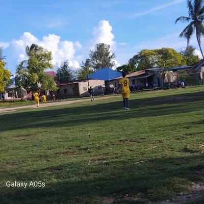 The image shows a soccer game in progress on a grassy field in Nungwi, Tanzania. The location is identified as -5.726 latitude, 39.301 longitude. There are five players in yellow jerseys visible, with one player in the foreground wearing the number 9 on his back. The player is walking away from the camera, and there are other players in the background. Several spectators can be seen sitting on the sidelines. There are several buildings in the background with thatched roofs and palm trees on the periphery of the field. The sky is bright blue with puffy white clouds. The ground is green grass with a few patches of dirt. The image was taken with a Samsung Galaxy A05s.
