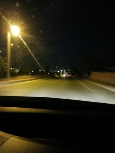 This is a first-person perspective, night-time shot from inside a vehicle traveling on a road. The scene is illuminated by streetlights, one of which is prominently visible on the left side of the image, casting a bright glow and lens flare. The road ahead appears to be asphalt and is lit by the vehicle's headlights, creating a bright path. Distant lights suggest a populated area or town. On the right side of the road, a low, textured wall is visible. The overall atmosphere is dark and somewhat hazy, with small dust particles or water droplets on the windshield creating small bokeh effects. The time of day is clearly night. The location is Bahşılı Belediyesi, Turkey, which provides context for the urban or semi-urban setting illuminated by streetlights. There is no visible text in the image.