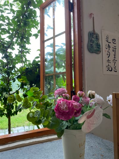 An open wooden window overlooks a lush green outdoor scene, possibly in Dujiangyan City, China, indicated by an artistic fan on the wall with Chinese calligraphy. Inside, a bouquet of artificial flowers, predominantly in shades of pink and white, sits in a cream-colored pot with "FLOW GARDEN" printed on it. The flowers are arranged with various green foliage. The window frame is visible, as is a portion of the wall with Chinese script and an ornamental fan. The weather outside appears to be overcast, with visible raindrops on the windowpane, suggesting a rainy day. The scene has a peaceful and artistic ambiance.