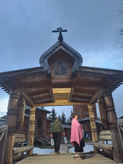 The image shows a wooden architectural structure, possibly an entrance or gate, with a cross at the top, adorned with an icon and intricate wood carvings. Two people are standing to the side of the structure: one wearing a pink shawl and black pants, the other in a green jacket and black pants. Both are holding water bottles. In the background, there are additional wooden structures suggesting a complex of buildings. This is located in the city of Vzglyadnevo, Russia.