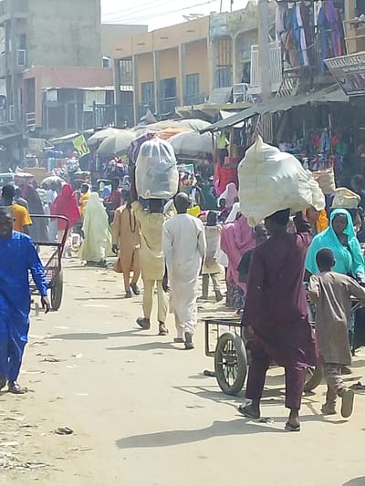 This image depicts a bustling outdoor market street in Fagge D1, Nigeria, under bright daylight conditions. The ground consists of an unpaved, dusty surface with visible debris. In the background, several multi-story concrete buildings with weathered facades are visible, alongside numerous market stalls covered by various awnings and umbrellas. Merchandise, including clothing, is displayed on and around these stalls.
Numerous individuals, primarily adults and some children, populate the scene, walking in various directions. They are dressed in diverse traditional West African attire, featuring colors such as blue, yellow, white, red, pink, brown, maroon, and aqua.
Specific individuals and objects include:
* In the mid-ground, two men are walking away from the viewer. Each carries a large, irregularly shaped bundle, wrapped in light-colored material (possibly white fabric or plastic), balanced on their heads. The man on the left wears a light brown robe, and the man on the right wears a white robe.
* In the left foreground, a man wearing a blue robe walks towards the viewer, pushing a dark-colored two-wheeled utility cart.
* In the right foreground, a man in a maroon robe walks away from the viewer, pushing a two-wheeled utility cart. A child in a light brown robe walks immediately to his left, also moving away from the viewer.
Long shadows cast by individuals and objects indicate a high sun angle. The scene conveys active commerce and pedestrian movement.