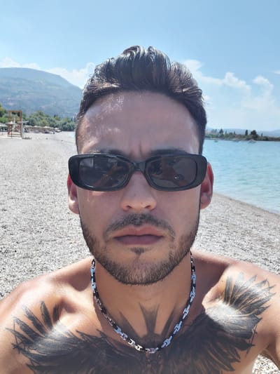 A man is taking a selfie on a pebble beach. The man is wearing black sunglasses, a silver chain necklace, and has tattoos on his chest. The background shows a body of water, mountains, and a clear blue sky with some clouds. This location is Municipal Unit of Diakopto, Greece.