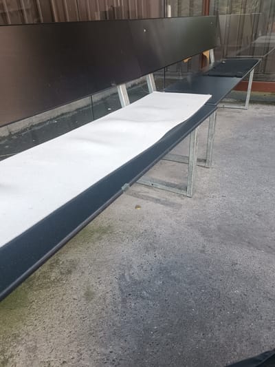 The image shows an empty outdoor bench with a white cushion on the seat. The bench has a black wooden seat and backrest, supported by a silver metal frame. The bench is placed on a concrete surface, with a building with glass windows in the background. The scene appears to be outdoors, possibly in a public space or a commercial area. There is no activity or event taking place, as the bench is unoccupied. The time of day and weather are not explicitly indicated, but the lighting suggests it might be daytime. There is no visible text in the image. The overall impression is one of stillness and a lack of human presence.