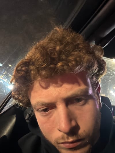 A man is captured in a close-up photograph, likely taken from an unusual angle or subsequently rotated. He has curly, light brown hair and a light skin tone. His facial features include visible stubble, and his eyes are open. He is wearing a dark-colored hooded garment. The background is dark, suggesting an interior setting, possibly a car, with a glimpse of indistinct lights visible through what seems to be a window. The location in Fort Lee, United States of America, cannot be confirmed with the given visual information.