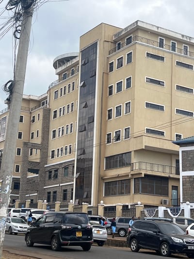 The image depicts a street scene in Rware ward, Kenya, featuring a tall, light-beige building with many windows. The building has multiple stories with balconies and dark-tinted windows, as well as some windows with white frames. A metal railing with a decorative pattern is visible on some of the balconies. Adjacent to the building, there are several parked cars, including a dark SUV with a license plate that appears to read "KDI 972E," a silver car, and a dark van. Some people are visible near the cars, seemingly going about their day. A utility pole with a dense network of wires is prominent in the foreground, suggesting an urban or semi-urban environment. The sky appears overcast, indicating a cloudy day. The scene captures a typical moment in a developed area, with architecture, vehicles, and infrastructure present.